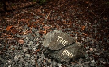 A rock with the words "THE END" painted on it, resting on gravel and surrounded by fallen leaves and twigs.