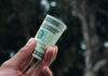 A close-up of a hand holding a rolled-up U.S. twenty-dollar bill, with a blurred natural background of trees and soft light.